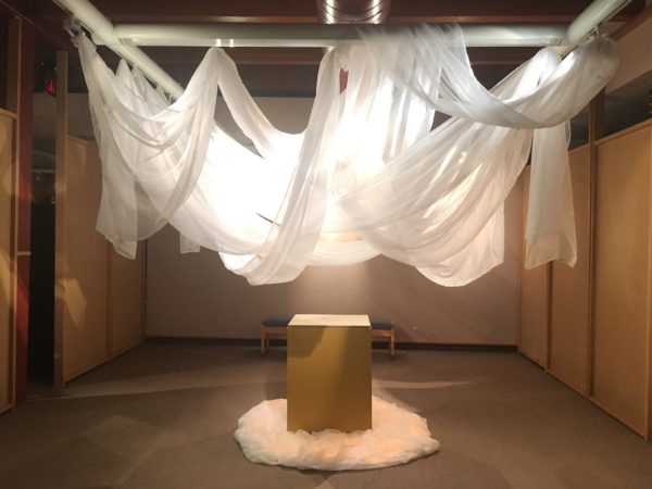 A brown box centered under white flowing cloth suspended in the air. A light is eminating from cloth.